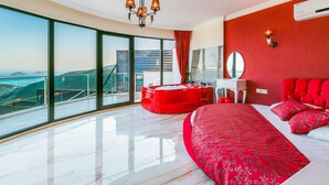 Un dormitorio moderno con una cama grande, un sillón rojo, un sofá rojo, un ventanal con vistas al paisaje y una araña.