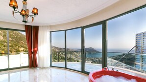 Amplia habitación con un ventanal que ofrece vistas panorámicas de un paisaje costero, una bañera roja y una lámpara moderna.