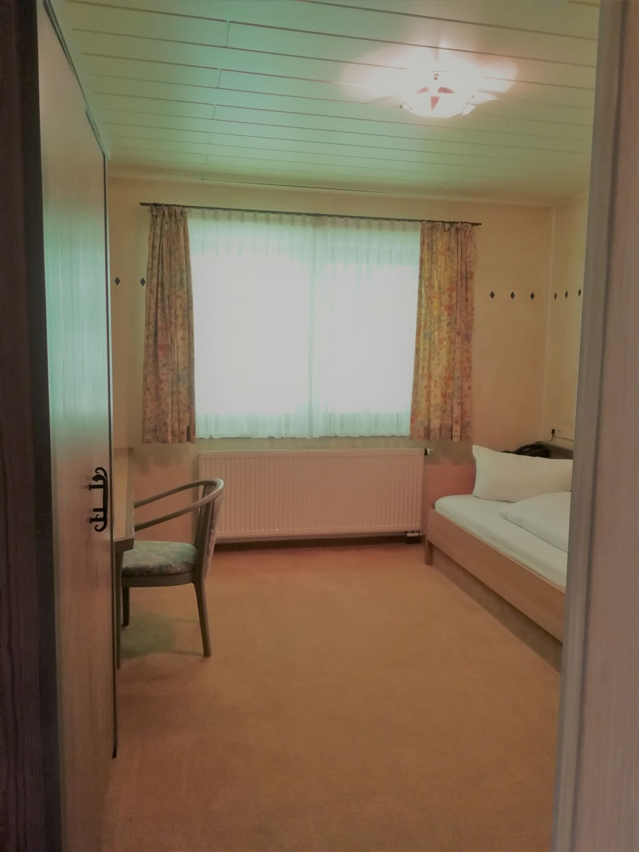 Eine kleine Einzimmerwohnung mit Bett, Stuhl, Heizkörper und einem fenster mit Vorhängen.