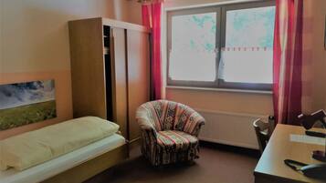A room with a bed, a chair, a desk with a computer, a radiator, and a window with curtains.