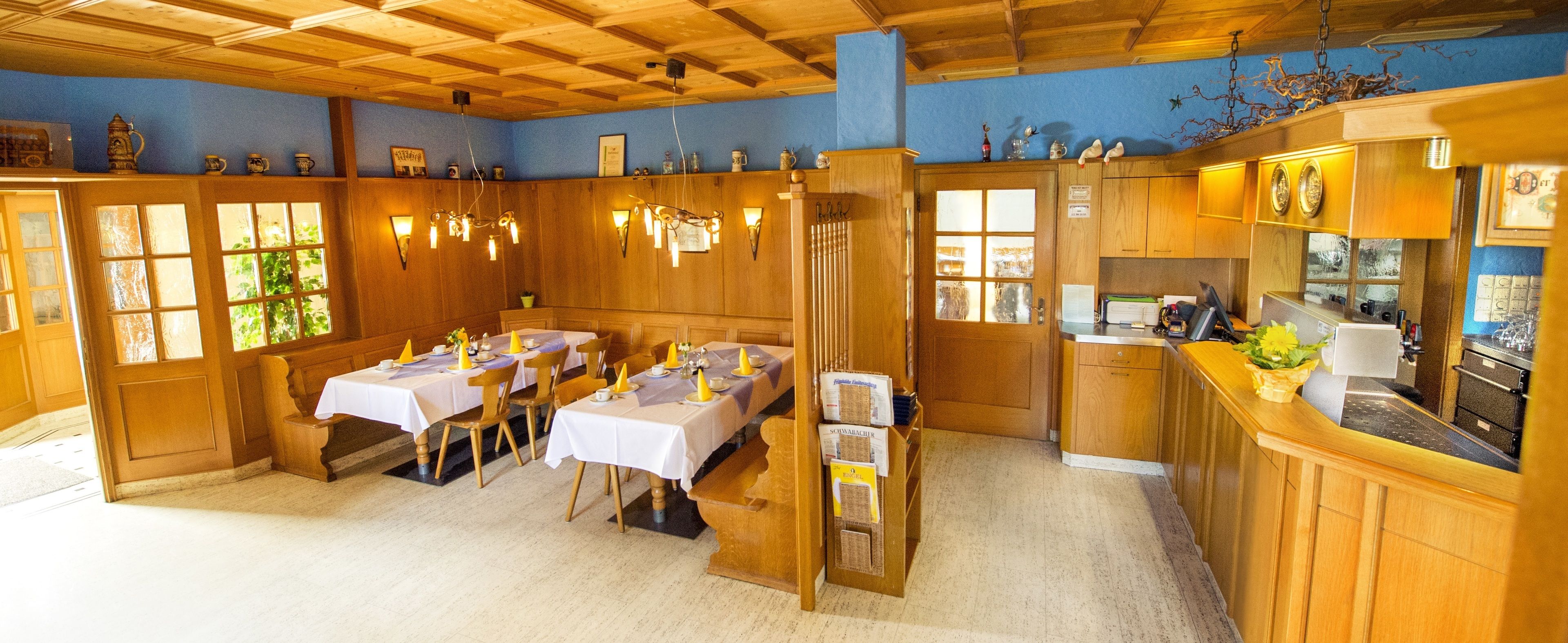 Een eethoek met houten tafels en stoelen, een keuken met een aanrecht en apparatuur, en een blauwe wand met decoratieve objecten.
