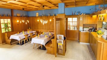 Een eethoek met houten tafels en stoelen, een keuken met een aanrecht en apparatuur, en een blauwe wand met decoratieve objecten.