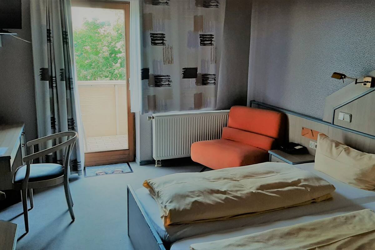 Une chambre d’hôtel équipée d’un lit, d’un bureau, d’une chaise, d’un radiateur et d’une fenêtre donnant sur un paysage verdoyant.