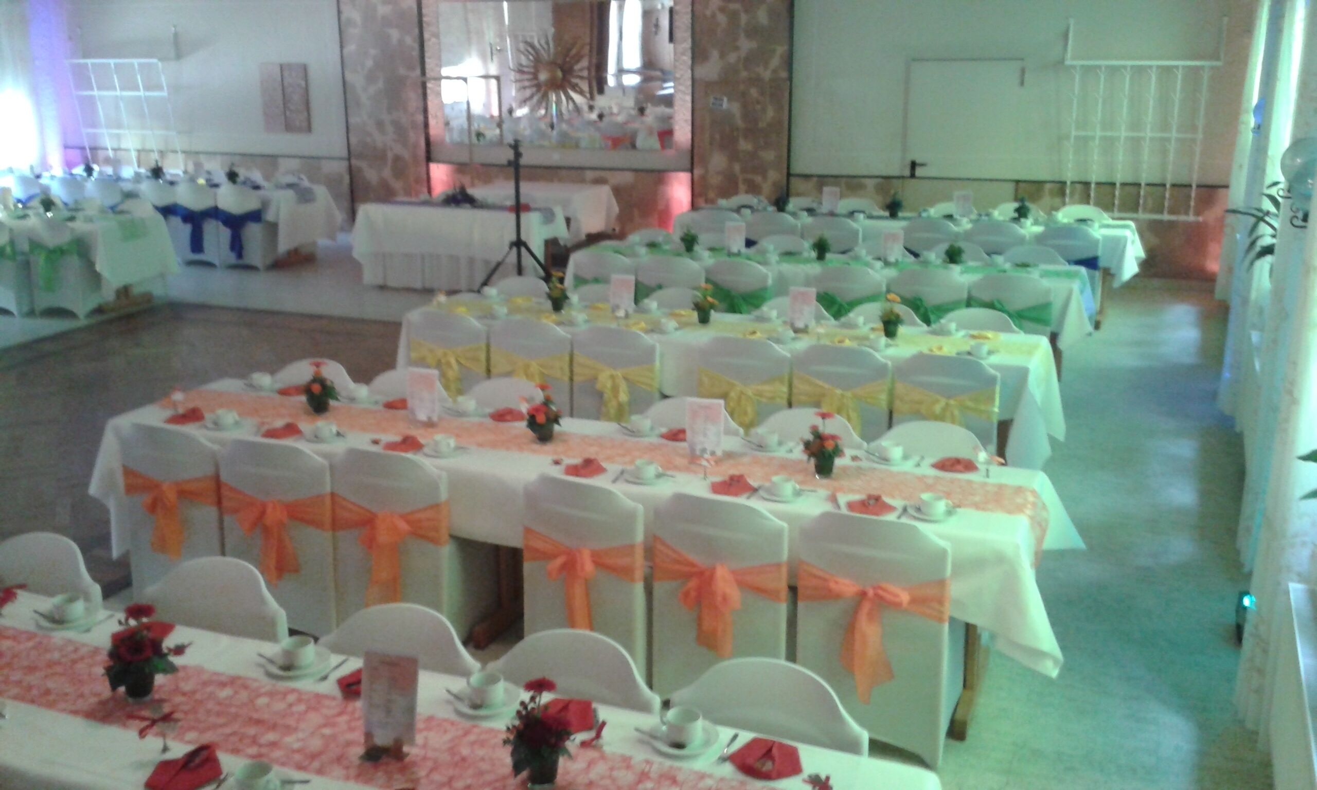 Un salón de banquetes con varias mesas dispuestas para un evento, cada una con manteles blancos y sillas adornadas con cintas naranjas.