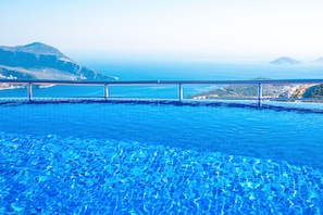 A clear view of a swimming pool with a metal railing, surrounded by a mountainous landscape and a distant view of the sea.