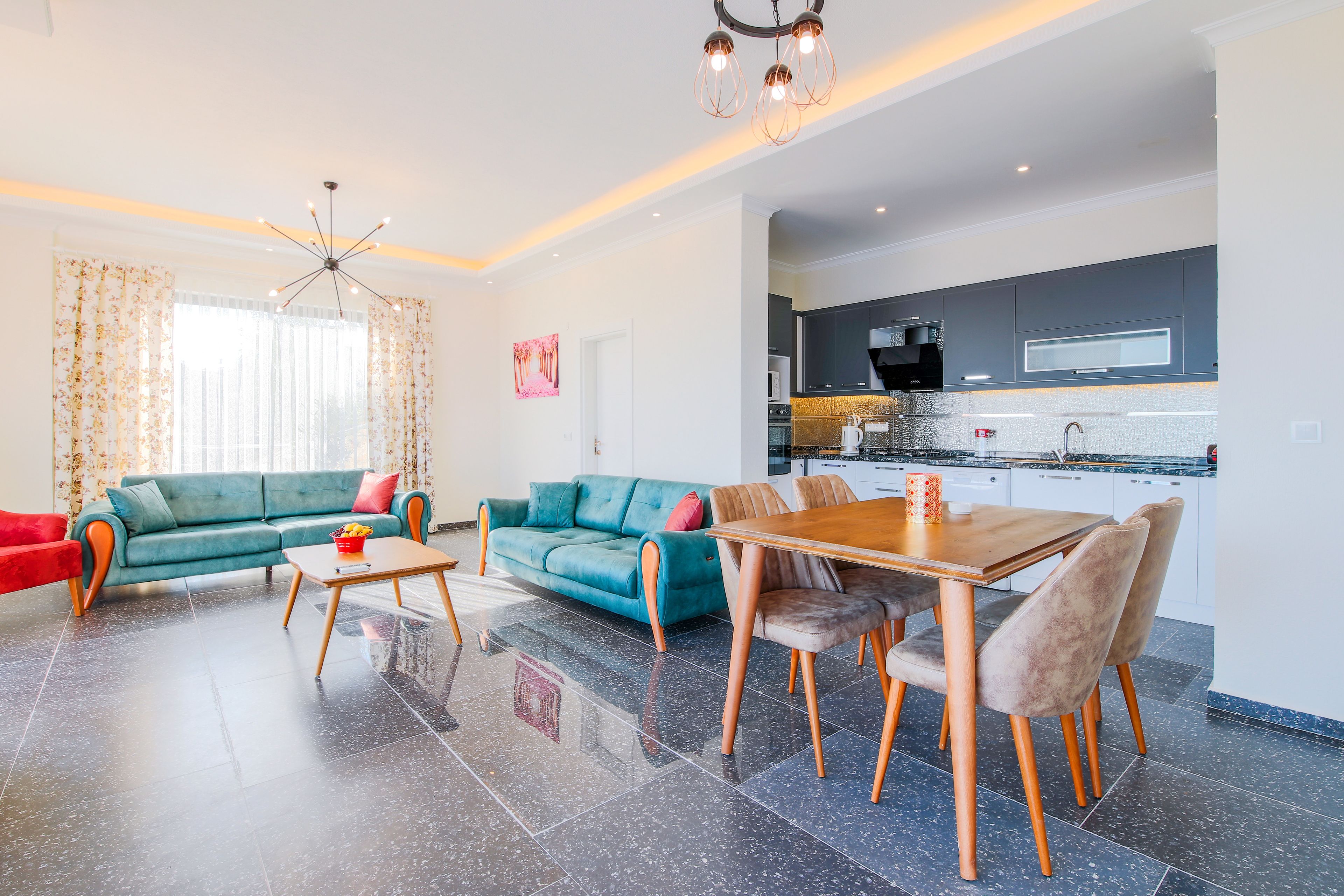 A modern living room with a sofa, a dining table, and chairs. There is a kitchen area with cabinets and appliances.