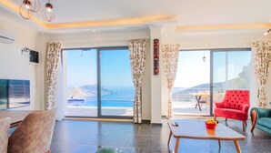 A spacious living area with a large sliding glass door leading to a balcony, a red armchair, a teal sofa, and a wooden coffee table. - Villa Gelincik-Hill Villas Kalkan (Kas)