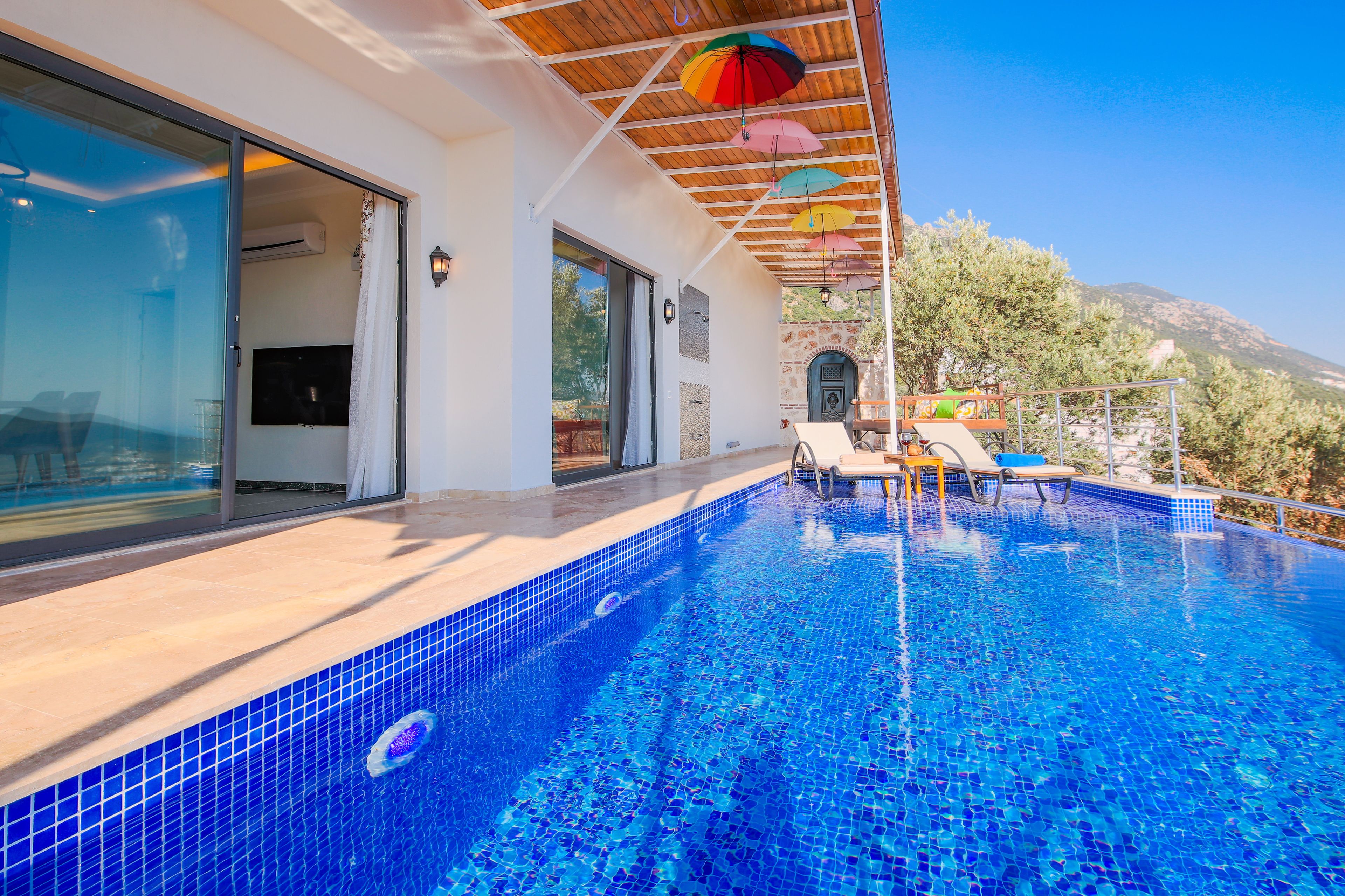Una piscina exterior moderna con suelo de azulejos azules, una zona cubierta con techo de madera y vistas a un paisaje montañoso.