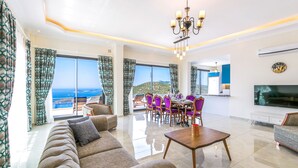 A spacious living room with a large sofa, dining table, and chairs, a TV, and a view of the sea.