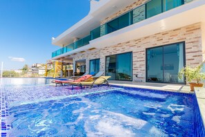 A modern poolside area with a clear blue pool, lounge chairs, and a contemporary building with large windows and a brick facade. - Villa Sato - Hill Villas Kalkan (Kas)