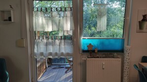 Una habitación con vistas a un balcón, una ventana con cortina de macramé y un pequeño estante de madera con objetos decorativos.