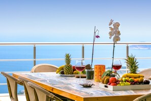 Una mesa con frutas, una copa de vino y una orquídea decorativa en un balcón con vistas al océano.