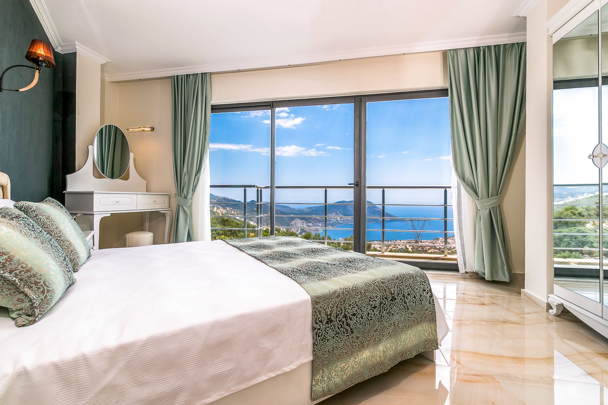 A bedroom with a large bed, a dresser, a mirror, and a view of the sea through large windows.