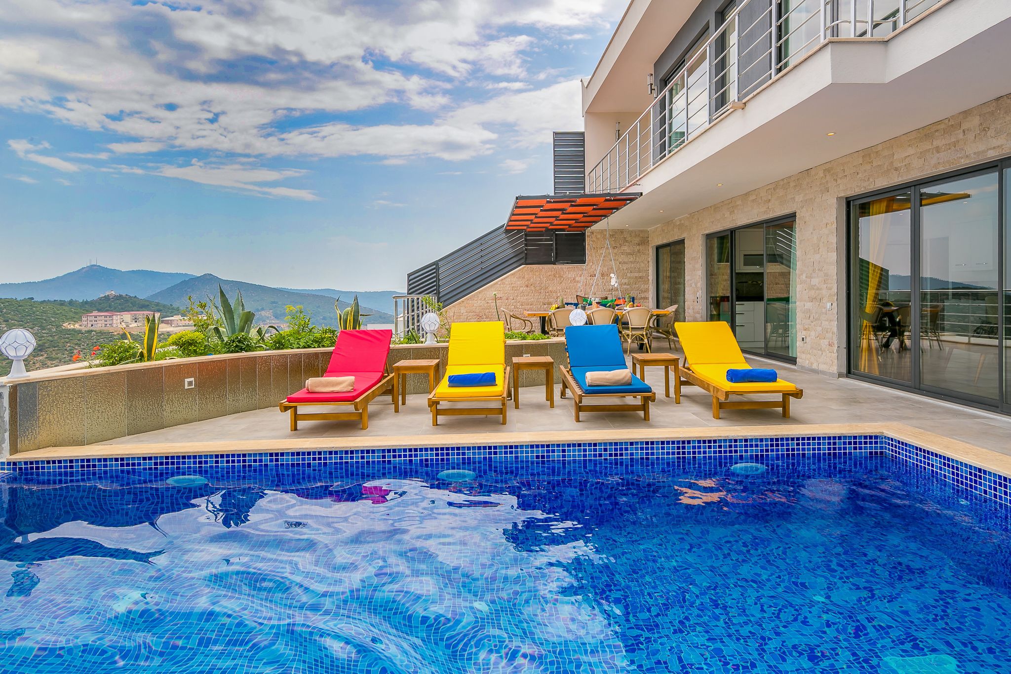 A rooftop pool area with a clear view of mountains, a modern building with large windows, and several colorful lounge chairs.