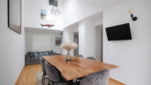 Un comedor moderno con una mesa de madera y sillas tapizadas en gris, un televisor de pantalla plana montado en la pared y una escultura decorativa de un pez.