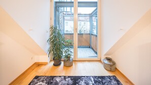 Una habitación con piso de madera, un tapete floral negro, una planta en maceta y un recipiente metálico.