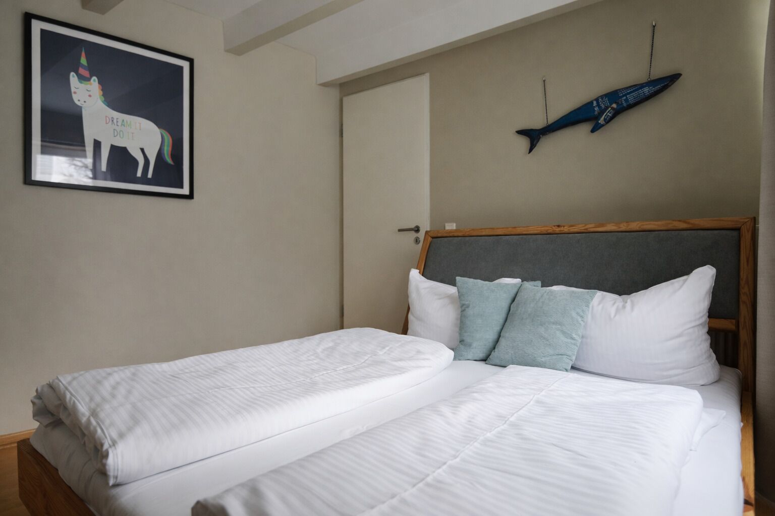Una cama doble con sábanas blancas, cabecera gris y dos almohadas. En la pared, hay un cuadro con un unicornio.