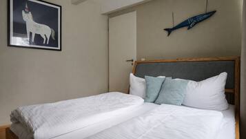 Una cama doble con sábanas blancas, cabecera gris y dos almohadas. En la pared, hay un cuadro con un unicornio.