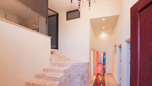 Un intérieur moderne avec un escalier en pierre menant à un deuxième étage, un couloir avec une porte rouge et une vue sur une pièce contenant un lit et une fenêtre.
