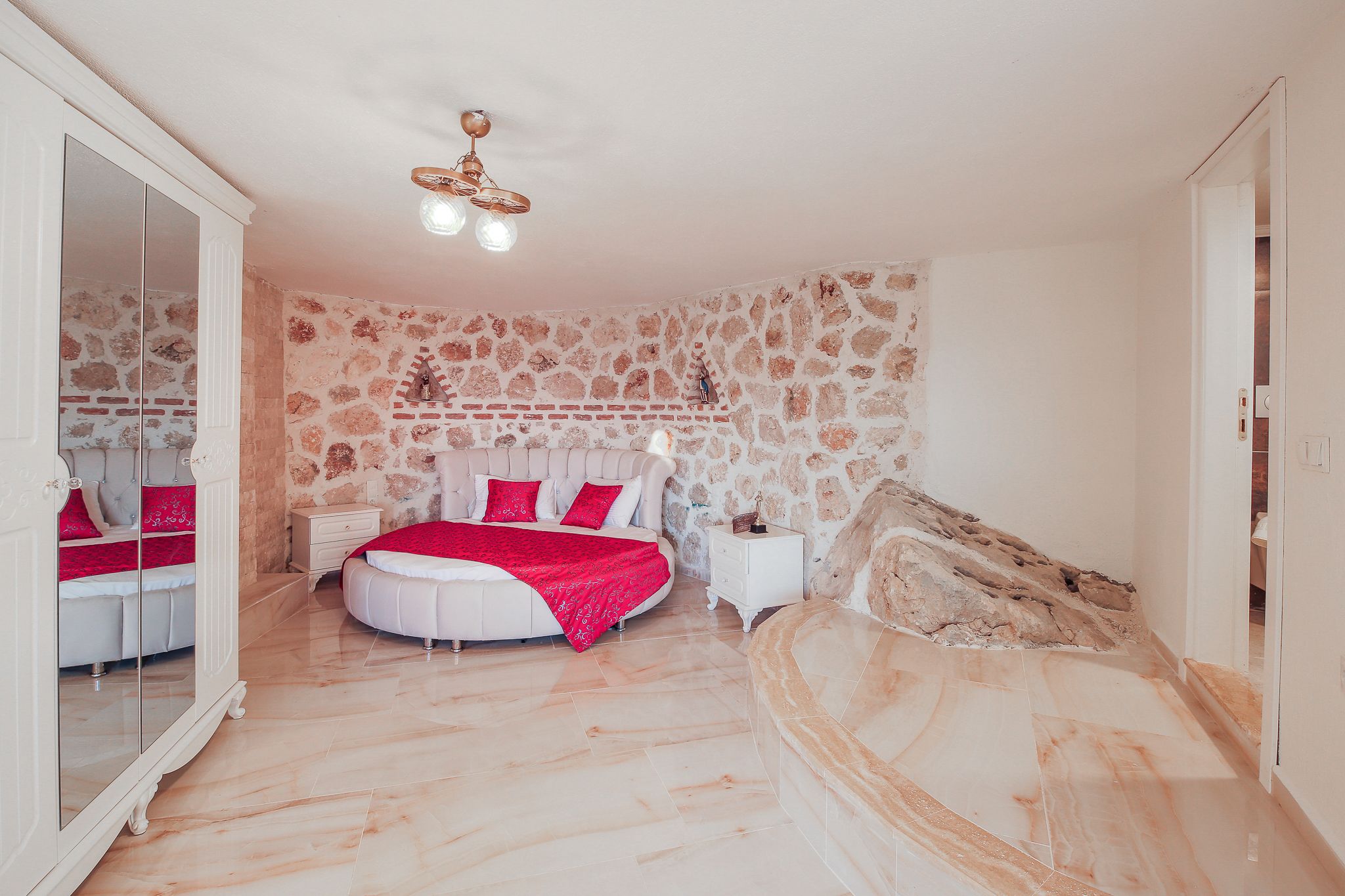 A bedroom with a stone wall, a round bed, a nightstand, and a mirror cabinet.