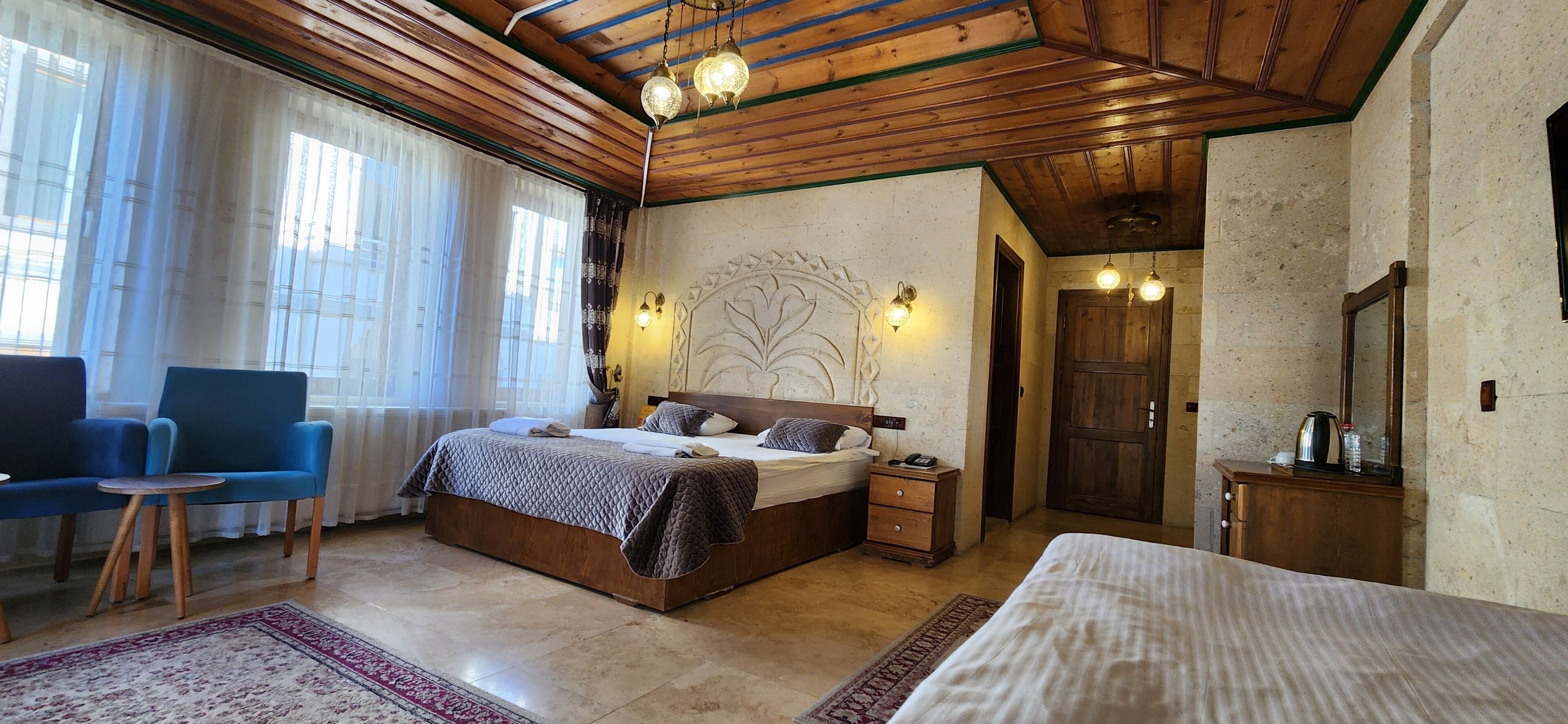 A bedroom with a wooden ceiling, a bed with a headboard, bedside tables, a chair, a small table, and a nightstand.