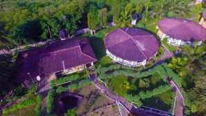 Vista aérea de un resort con varias construcciones, incluyendo una gran estructura circular con techo de tejas, rodeado de exuberante vegetación y palmeras.
