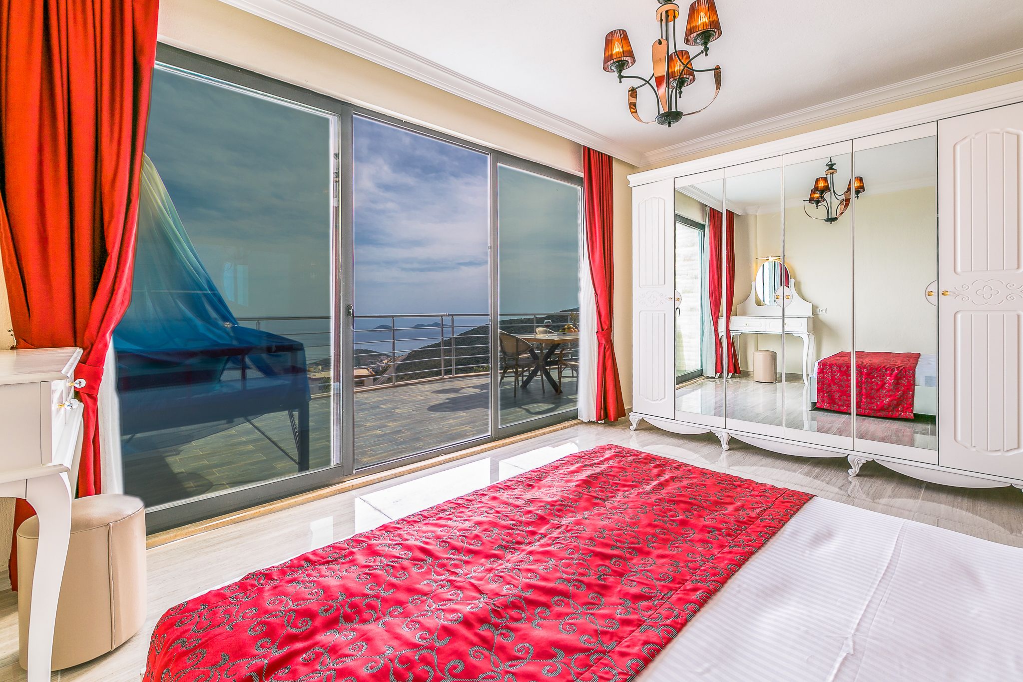 A bedroom with a large sliding glass door to a balcony, a bed with a red patterned bedspread, a white wardrobe, and a piano in the corner.