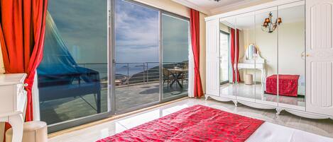 A bedroom with a large sliding glass door to a balcony, a bed with a red patterned bedspread, a white wardrobe, and a piano in the corner.