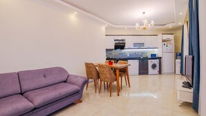 A modern living room with a purple sofa, a dining table with four chairs, a washing machine, and a refrigerator in the background.