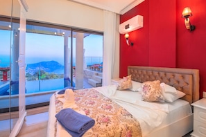 A hotel room with a large bed, a red accent wall, and a balcony with a view of the sea and mountains.