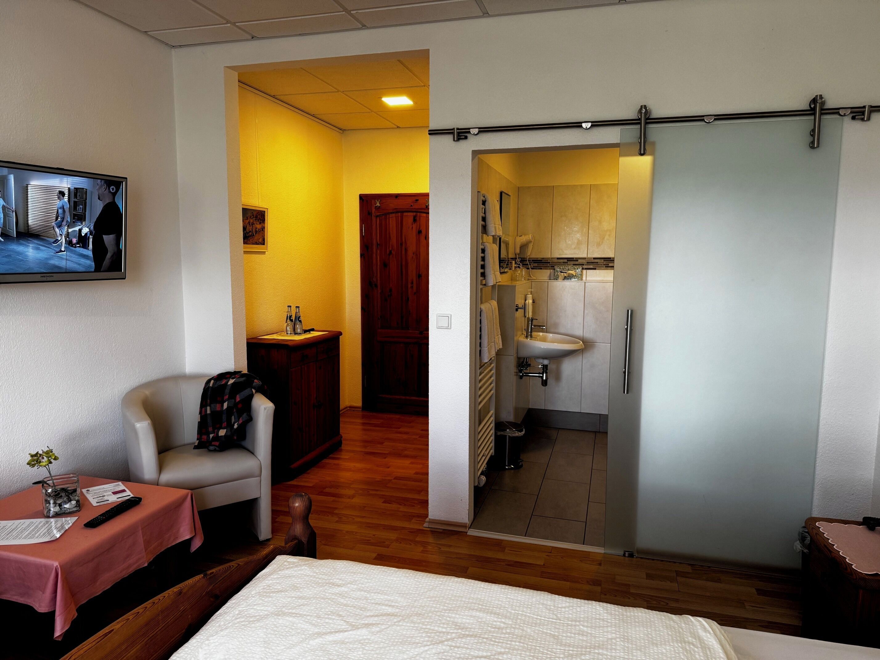 A hotel room with a bed, a small table with a vase and remote control, a wooden chair, a wooden cabinet, and a bathroom with a sink and mirror.