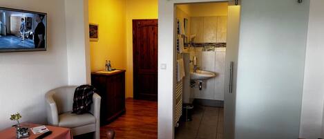 A hotel room with a bed, a small table with a vase and remote control, a wooden chair, a wooden cabinet, and a bathroom with a sink and mirror.
