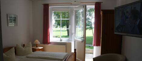 A hotel room with a double bed, a chair, a table with bottles, a TV, and a window with curtains.