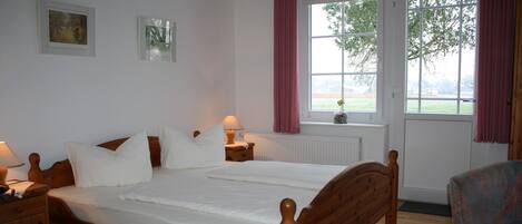 A wooden bed in a room with a window, a chair, and a nightstand with a lamp.