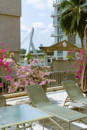 Un balcón con sillones de descanso, una palmera y un puente al fondo.