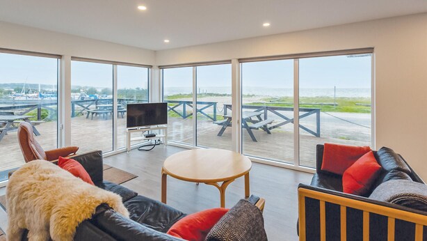 Un salón moderno con un televisor grande, asientos cómodos y vistas a la playa y barcos a través de amplias ventanas.