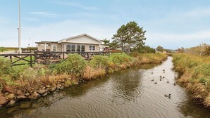 Un edificio a orillas del río con un muelle, rodeado de vegetación natural y patos en el agua.