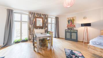 Ein Zimmer mit Holzboden, einem Esstisch mit Stühlen, einem Bett, einer Lampe, einem Schrank und einem großen Fenster mit Vorhängen.