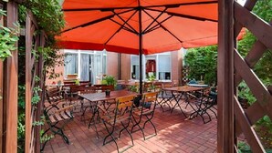 Een terras buiten met bakstenen vloer, houten tafels en stoelen, en een grote oranje parasol voor schaduw.