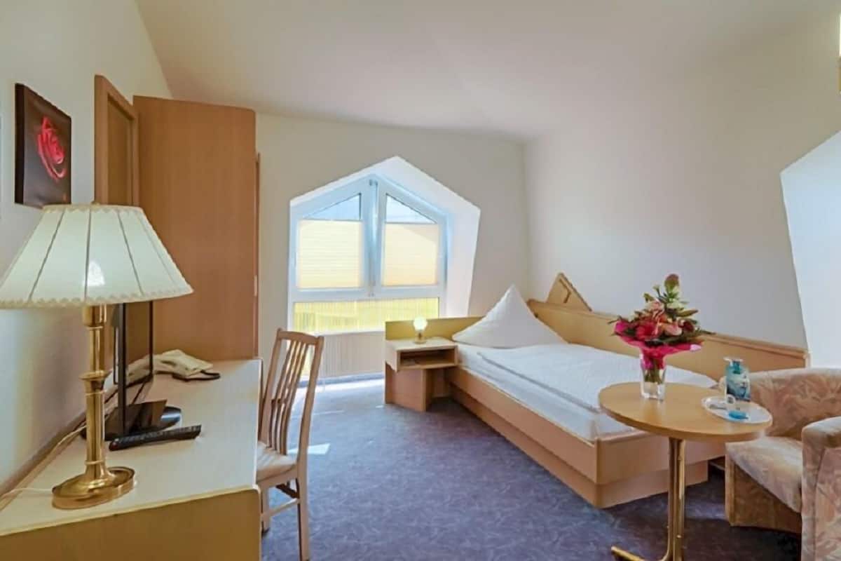 Ein Hotelzimmer mit Bett, Schreibtisch, Stuhl, einem kleinen Tisch mit Blumenvase und einem Fenster mit Jalousien.