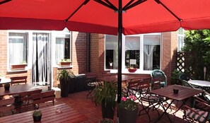 Une terrasse extérieure avec des tables et des bancs en bois, entourée de plantes en pot et agrémentée d’un parasol rouge.