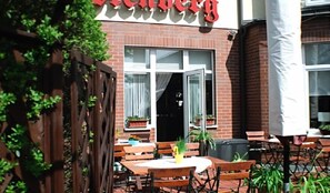 An outdoor seating area with wooden tables and chairs, potted plants, and a brick building with a red awning.