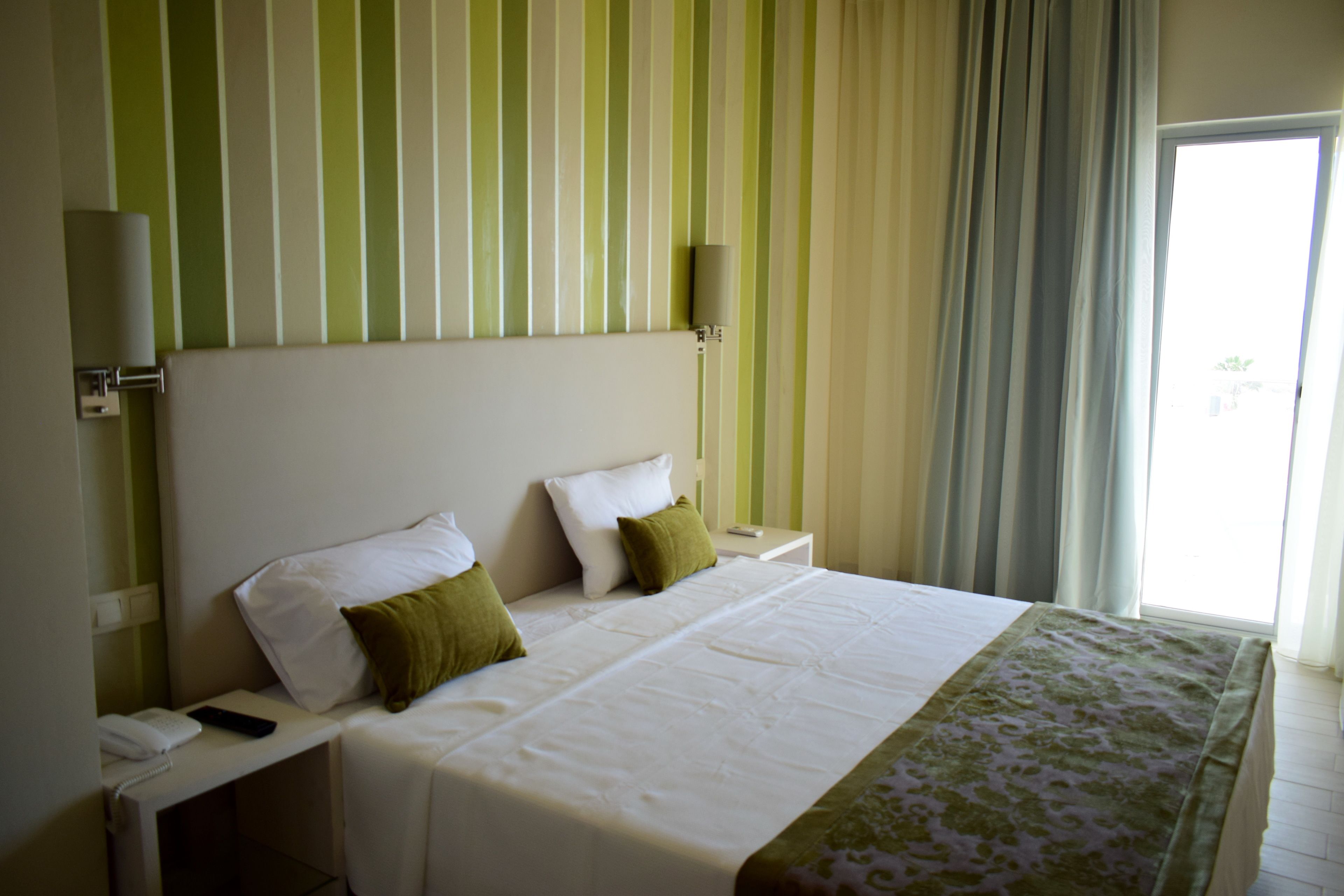 Habitación de hotel con una cama grande, dos almohadas, una mesita de noche con lámpara y una ventana con cortinas.