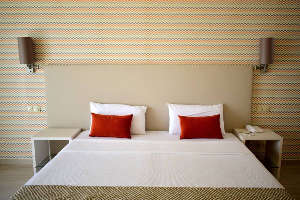 A hotel room with a large bed, two red pillows, a beige headboard, and patterned wallpaper.