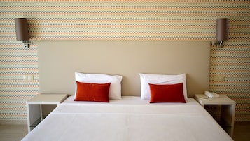 Quarto de hotel com uma cama grande, duas almofadas vermelhas, cabeceira bege e papel de parede estampado.
