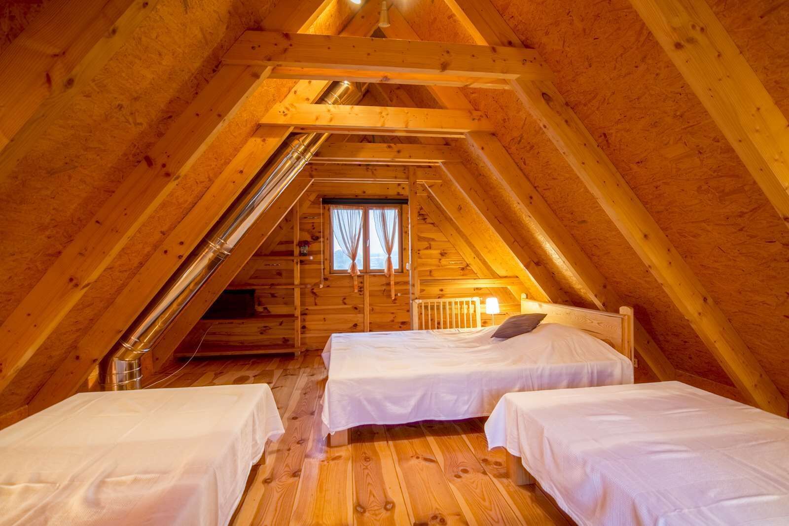 Een gezellige slaapkamer onder de dakpannen met twee bedden, houten balken en een raam met gordijnen.