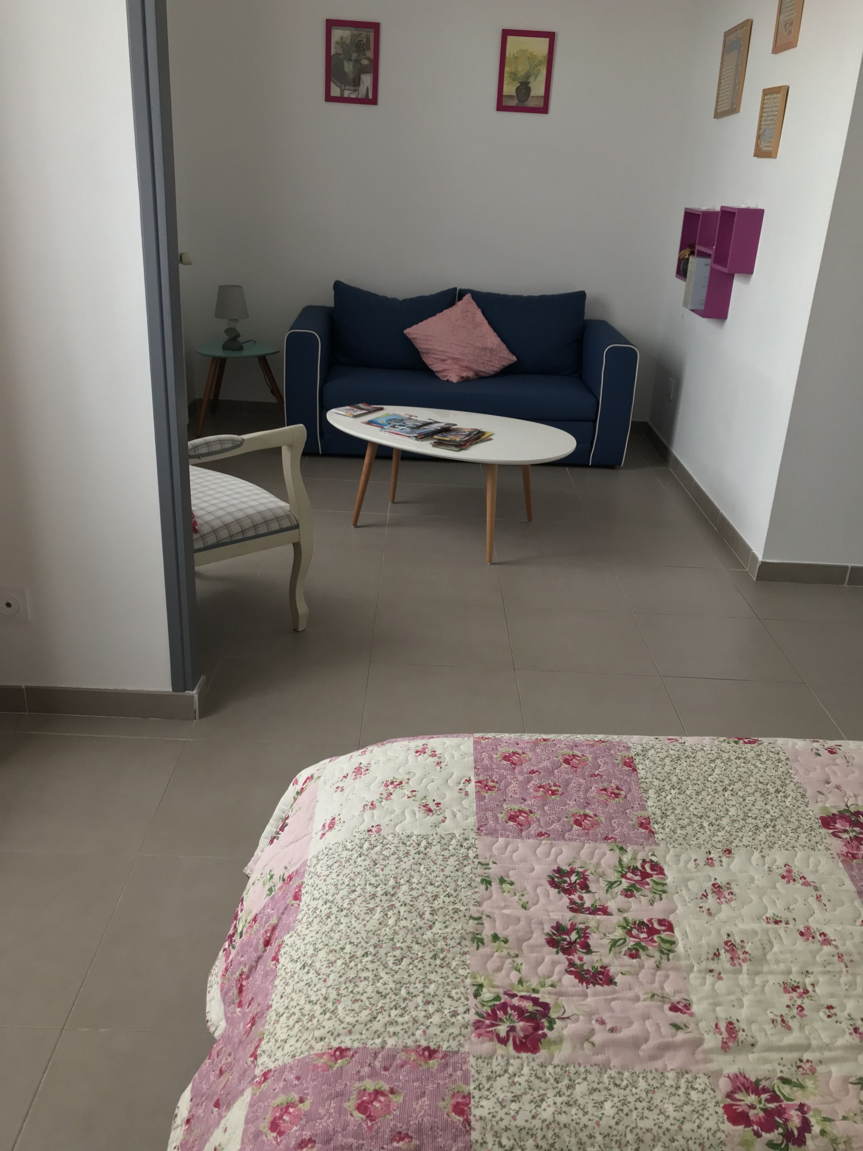 Ett rum med en blå soffa, ett vitt soffbord, ett blommigt sängöverkast och ett litet bord med en lampa.