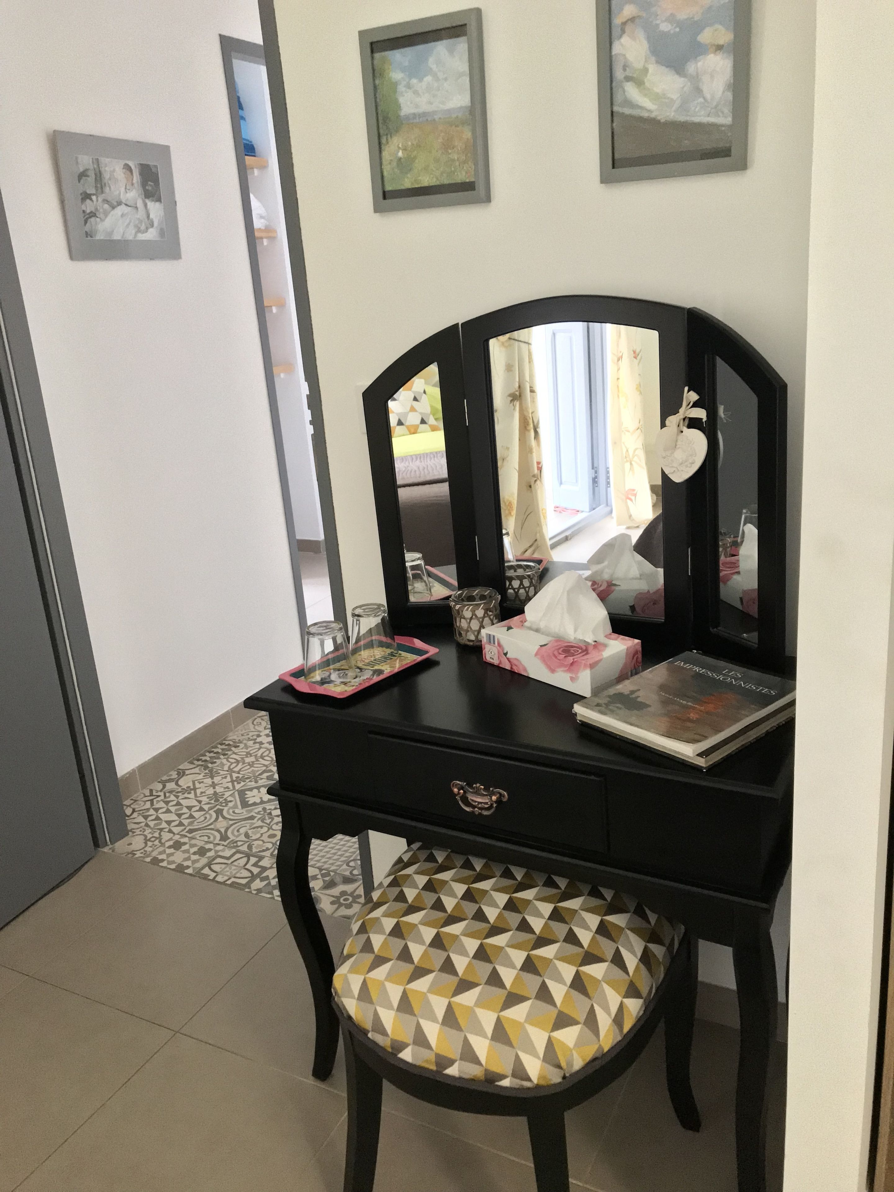 Une pièce avec une coiffeuse noire, une chaise à motifs et un miroir. Les murs sont ornés de tableaux encadrés et il y a une fenêtre avec des rideaux.