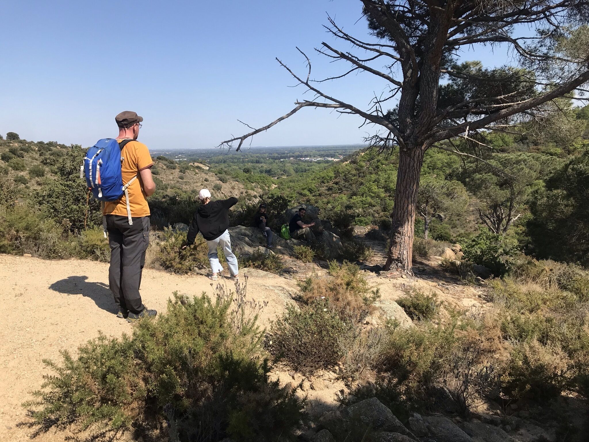Un excursionista con una mochila azul y otro excursionista con una chaqueta negra en un sendero de tierra con vegetación escasa y cielo despejado.