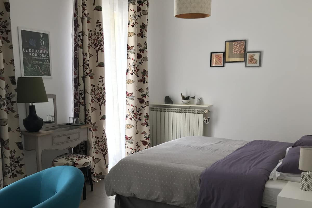 Un dormitorio con cama, una silla, una mesita de noche, una lámpara, un radiador y una pared decorada con cuadros.